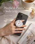 A beautifully styled commercial-style top-down photo featuring an open hand holding a black smartphone. The phone's back is fitted with a circular grip designed as a playful, illustrative clock face (showing numbers and cute icons). The collapsing base has white and pink stripes. Decorative background elements include pink tulips in a ribbed vase, a cup of espresso, a bottle of Zara perfume, a magazine (