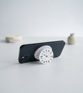 A black smartphone propped up horizontally on a table, using a pop-out grip with a playful, hand-drawn-style clock face. The clock dial shows numbers and small, cute illustrations (flowers, clouds, a heart, etc.). The collapsible base is white with soft pink stripes. In the soft-focus background are small pastel discs and a tiny vase.