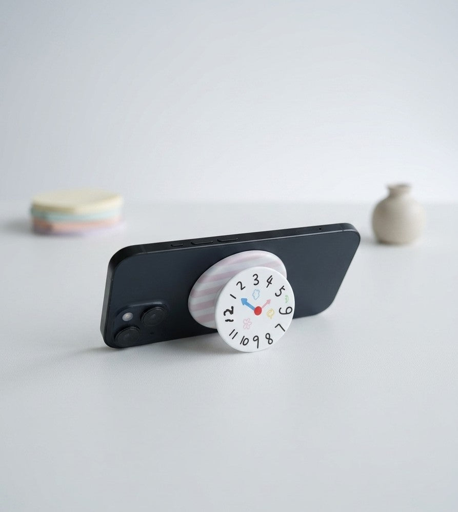 A black smartphone propped up horizontally on a table, using a pop-out grip with a playful, hand-drawn-style clock face. The clock dial shows numbers and small, cute illustrations (flowers, clouds, a heart, etc.). The collapsible base is white with soft pink stripes. In the soft-focus background are small pastel discs and a tiny vase.