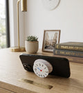 A black smartphone propped up horizontally on a wooden desk, standing on a pop-out grip designed as a playful, hand-drawn clock face. The collapsible base is white with pink stripes. The desk environment includes a large brass lamp, a potted succulent, a framed photo of a European street scene, and a stack of books with the top book titled 