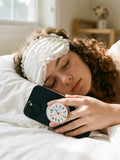 A close-up photograph of a woman with curly brown hair, wearing a white silk and lace sleeping mask with bows, resting in a comfortable white-sheet bed. Her hand holds up a dark smartphone, showing the circular grip with a hand-drawn clock design (numbers, heart, and cloud doodles). The collapsible base is white with pink stripes. In the soft-focus background are warm light and a wooden object.