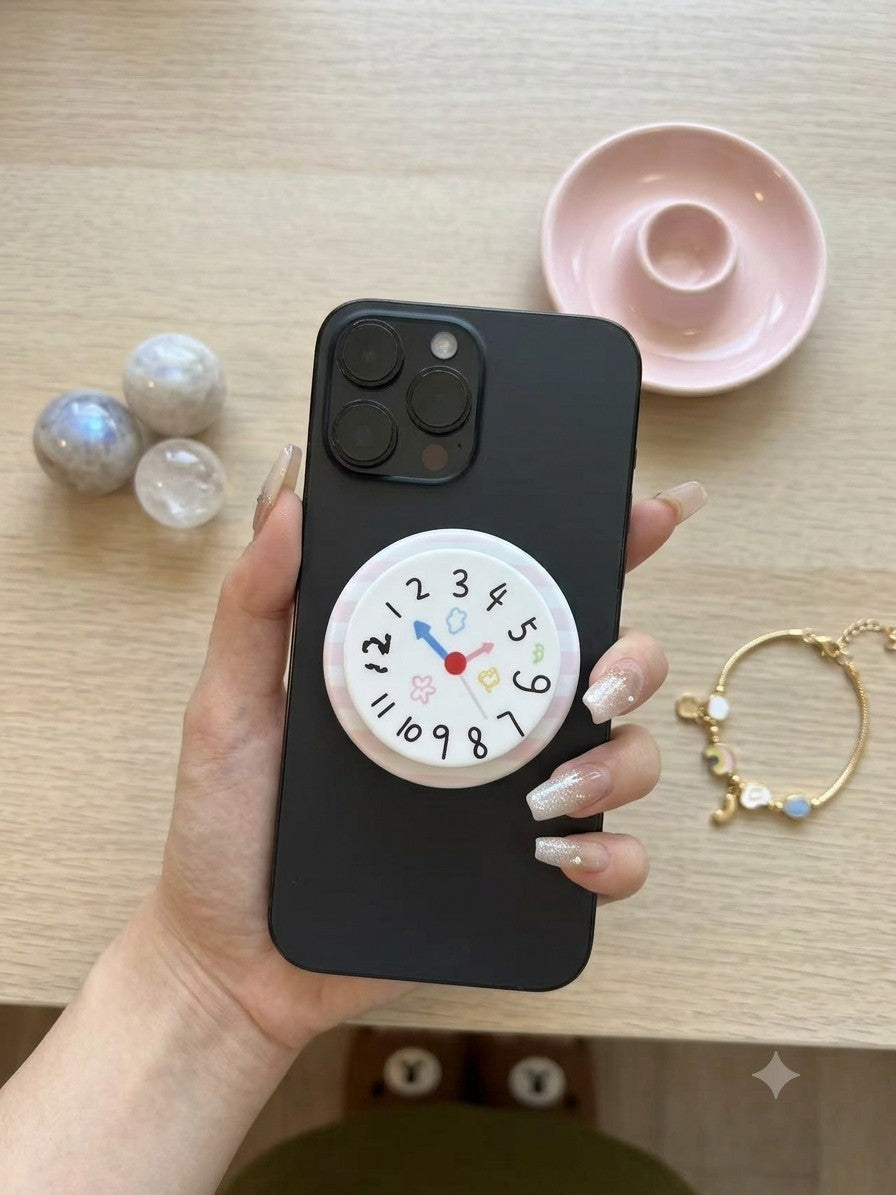 A top-down photograph on a light wooden table, showing a hand with elegant, nude and glitter-tipped nails, holding a dark smartphone. A large circular grip with a playful, illustrative clock face (showing numbers and tiny icons) is attached to the back. The collapsible base is white with soft pink stripes. The surrounding table features decorative items: a stack of marble coasters, a pink ring holder, a small porcelain object, and a delicate gold chain bracelet with pastel charms.