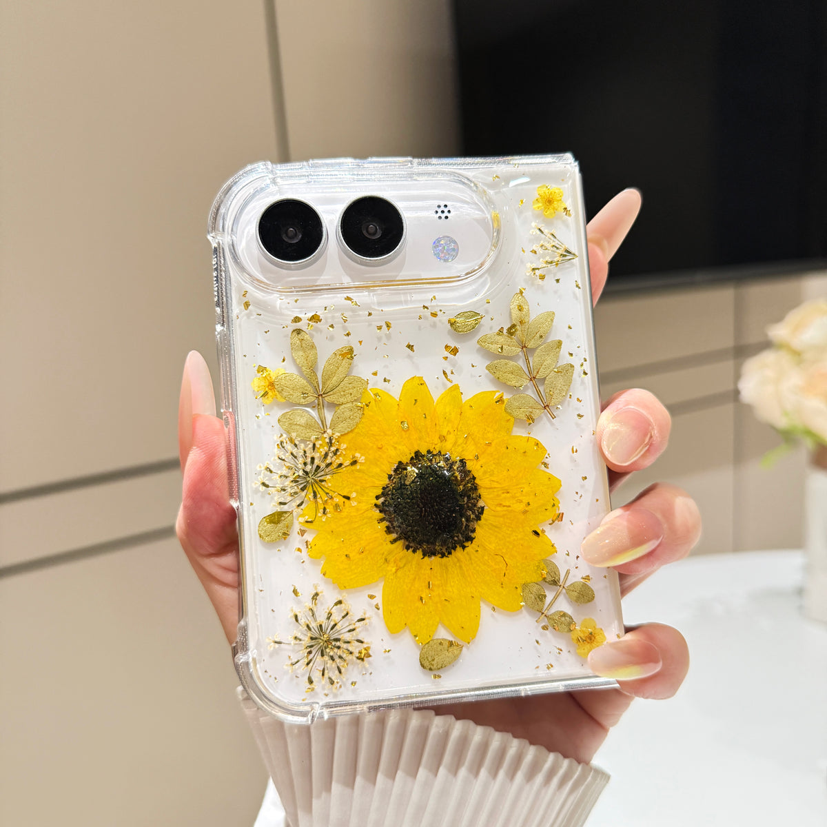 Back cover view of a hand holding the clear iPhone DUO case embedded with a real pressed yellow sunflower, green foliage, and gold foil flecks around the dual camera cutout.