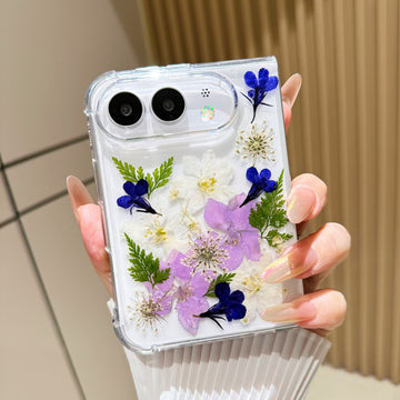 Back cover view of a hand holding the clear iPhone DUO case embedded with real pressed blue, purple, and white flowers and green fern sprigs around the dual camera cutout.