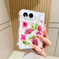 Back cover view of a hand holding the clear iPhone DUO case embedded with real pressed pink flowers, green leaves, and silver foil flecks around the dual camera cutout.