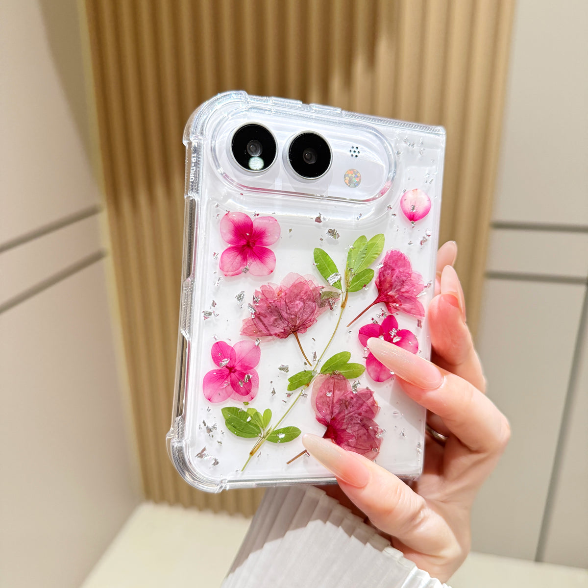 Back cover view of a hand holding the clear iPhone DUO case embedded with real pressed pink flowers, green leaves, and silver foil flecks around the dual camera cutout.