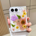Back cover view of a hand holding the clear iPhone DUO case embedded with real pressed pink tulips, yellow daisies, green fern fronds, and gold foil flecks around the dual camera cutout.