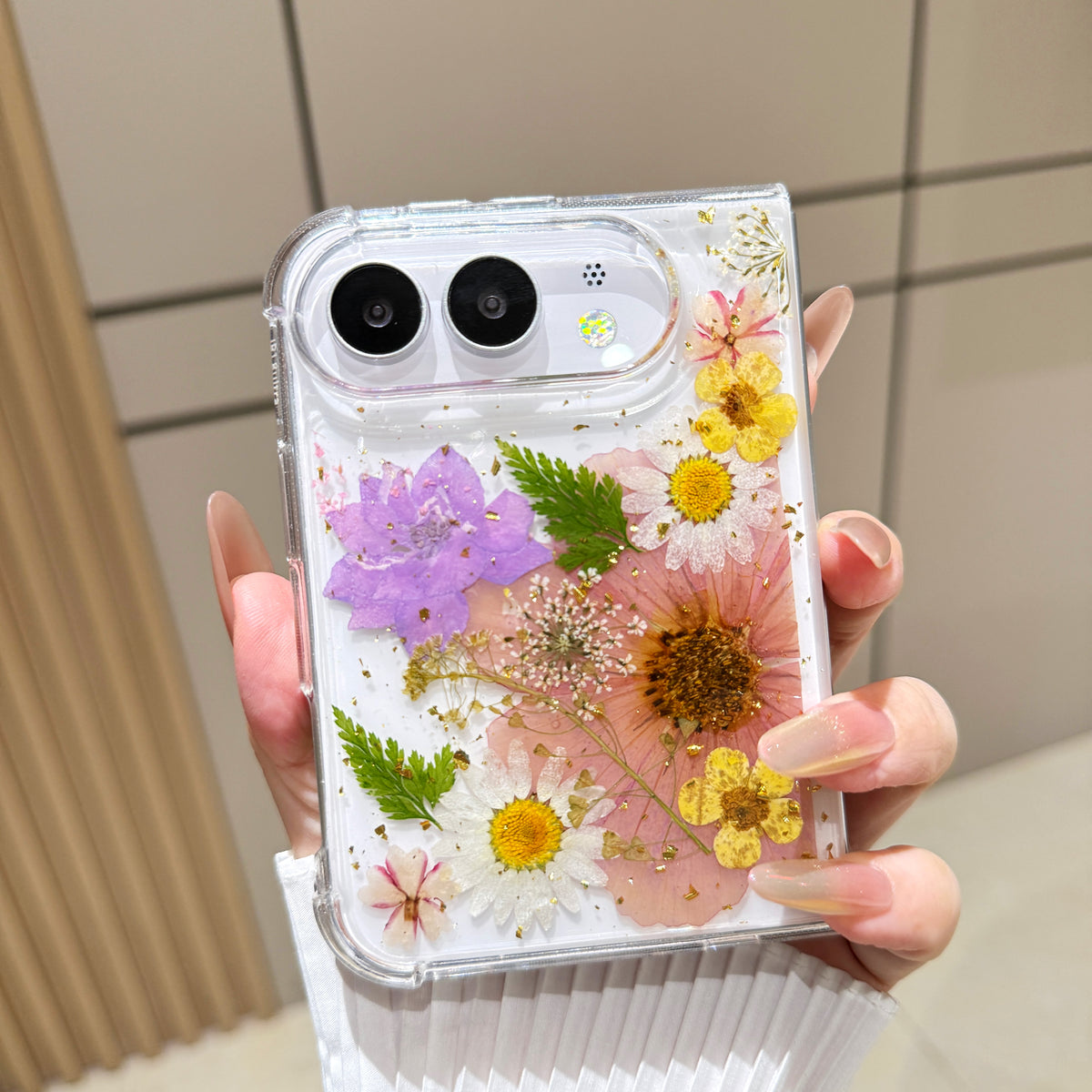 Back cover view of a hand holding the clear iPhone DUO case embedded with real pressed pink tulips, yellow daisies, green fern fronds, and gold foil flecks around the dual camera cutout.