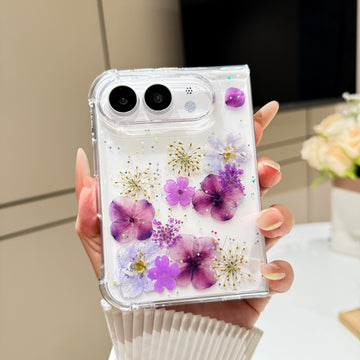 Back cover view of a hand holding the clear iPhone DUO case embedded with real pressed purple flowers, delicate botanical stems, and shimmer glitter around the dual camera cutout.