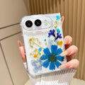 Back cover view of a hand holding the clear iPhone DUO case embedded with real pressed royal blue daisy flowers, yellow botanical sprigs, and fine shimmer glitter around the dual camera cutout.