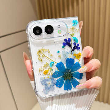 Back cover view of a hand holding the clear iPhone DUO case embedded with real pressed royal blue daisy flowers, yellow botanical sprigs, and fine shimmer glitter around the dual camera cutout.