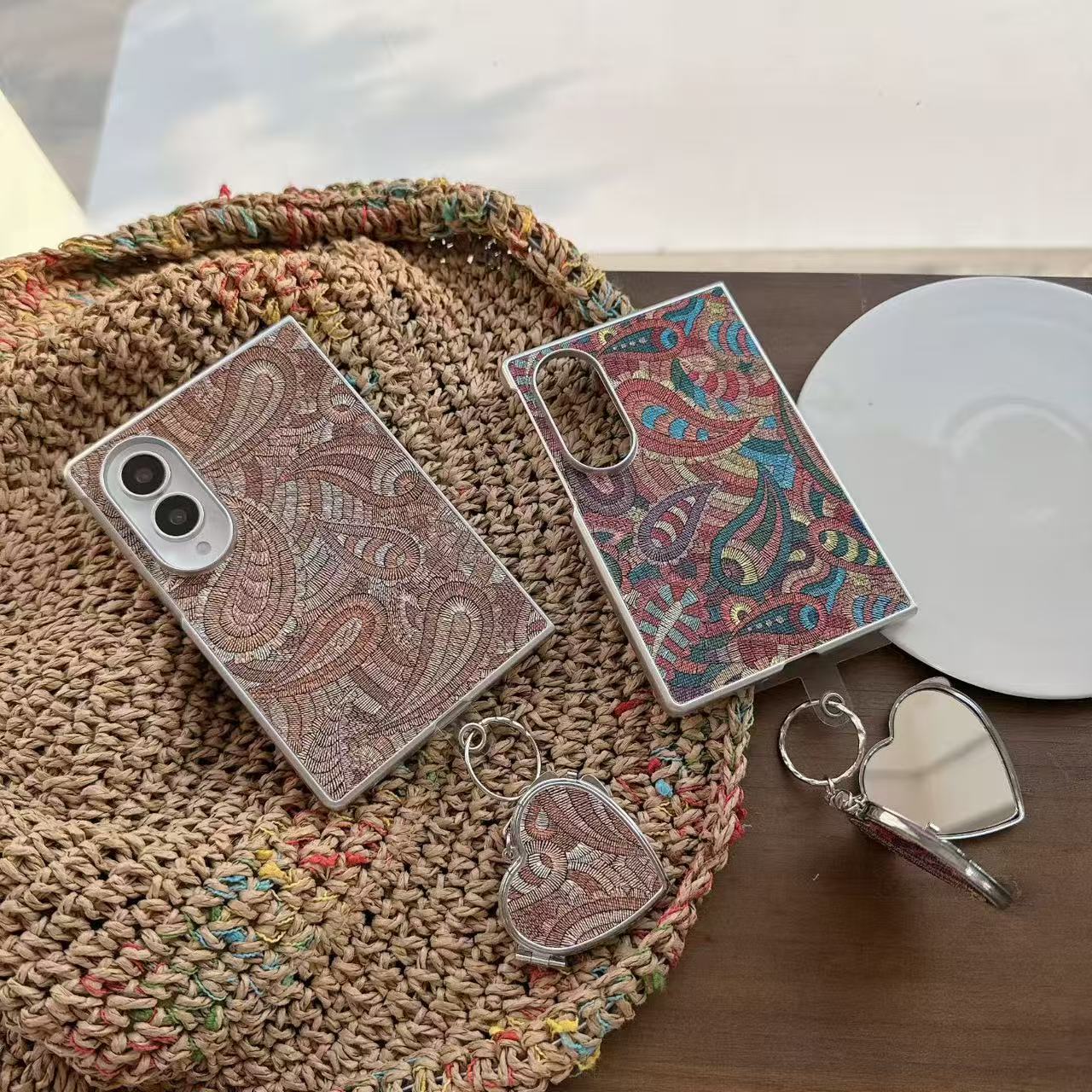Brown and multi-color paisley tapestry Samsung Galaxy Z Fold 8 cases resting on a woven straw tote bag with attached heart compact mirror charm.