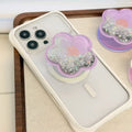 Lavender purple flower-shaped magnetic MagSafe phone grip attached to a white iPhone case placed on a wooden display tray with collapsible kickstand closed.