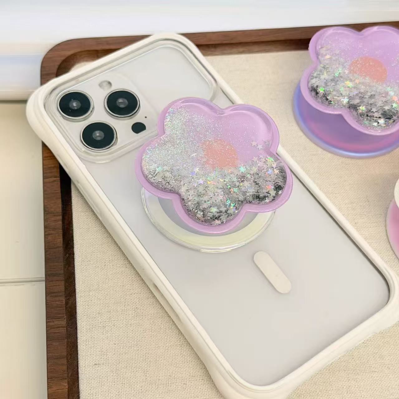 Lavender purple flower-shaped magnetic MagSafe phone grip attached to a white iPhone case placed on a wooden display tray with collapsible kickstand closed.