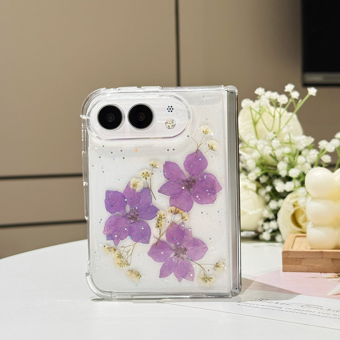 Side-by-side display of two iPhone DUO clear glitter cases showcasing the handmade real pressed flower options in elegant violet purple and soft blossom pink.