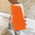 Close-up hand view of the vibrant orange dual-tone leather wallet case held against a magazine, highlighting the cream contrast panel and brass snap button.