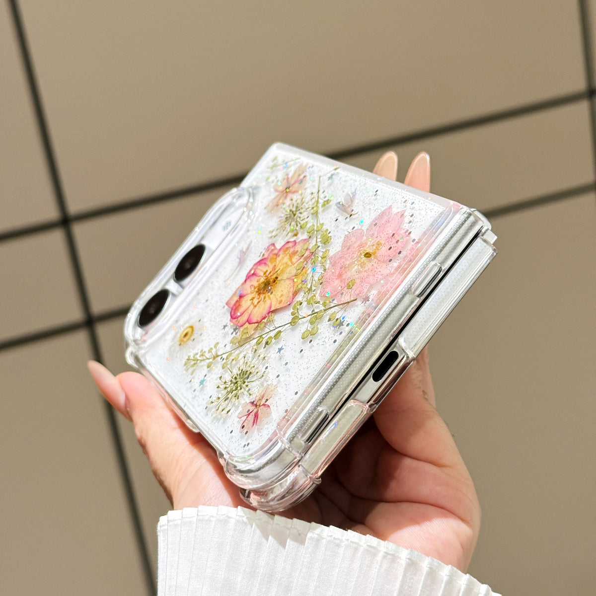 Close-up side view of a hand holding the clear iPhone DUO real pressed flower case at a bottom angle, showing the charging port cutout, precision hinge clearance, and embedded floral arrangement.