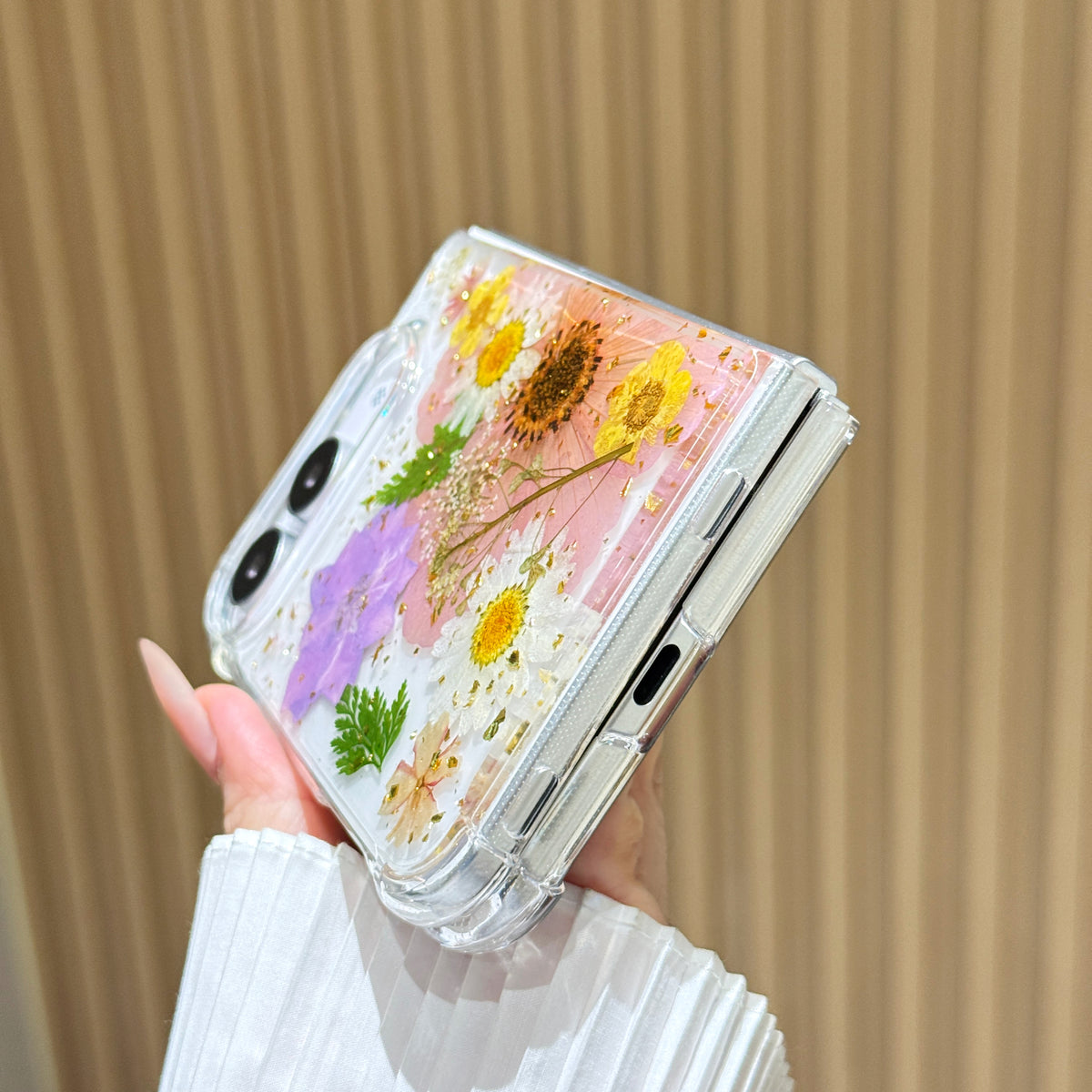 Close-up side view of a hand holding the clear iPhone DUO real pressed flower case at a bottom angle, showing the charging port cutout, precision hinge clearance, and embedded floral garden arrangement.