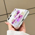 Close-up side view of a hand holding the clear iPhone DUO real pressed flower case at a bottom angle, showing the charging port cutout, precision hinge clearance, and embedded purple floral arrangement.