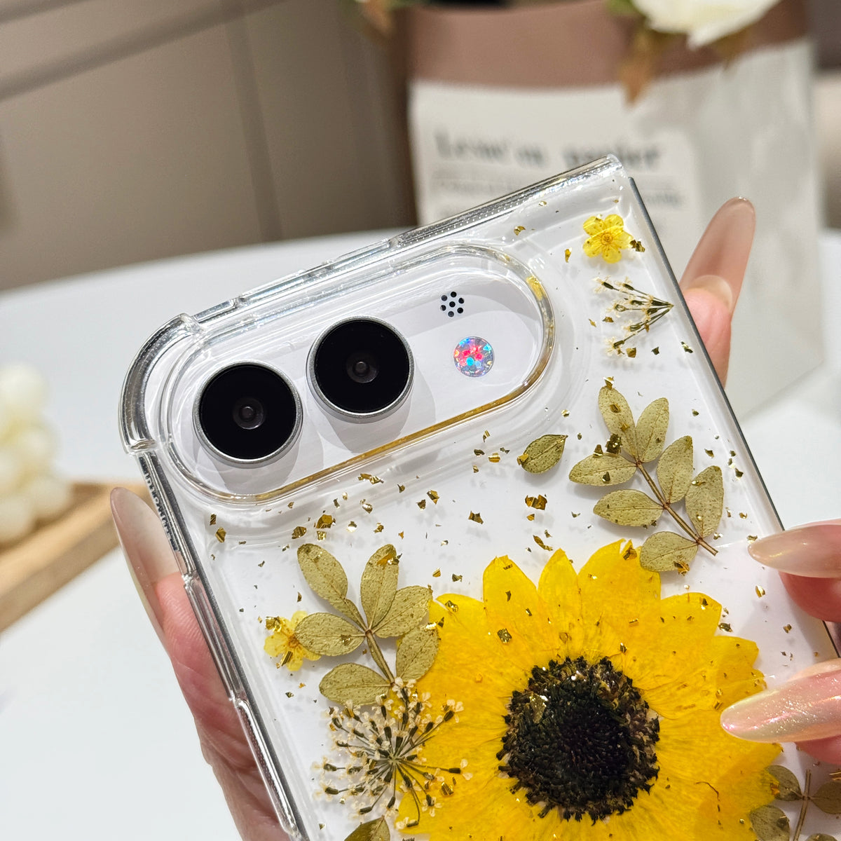 Close-up top-angle view of the clear iPhone DUO case embedded with a real pressed yellow sunflower, fine botanical leaves, and gold foil flakes surrounding the dual camera lens guard.
