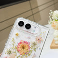 Close-up top-angle view of the clear iPhone DUO case embedded with real pressed dry flowers, fine botanical stems, and holographic star glitter surrounding the dual camera lens guard.