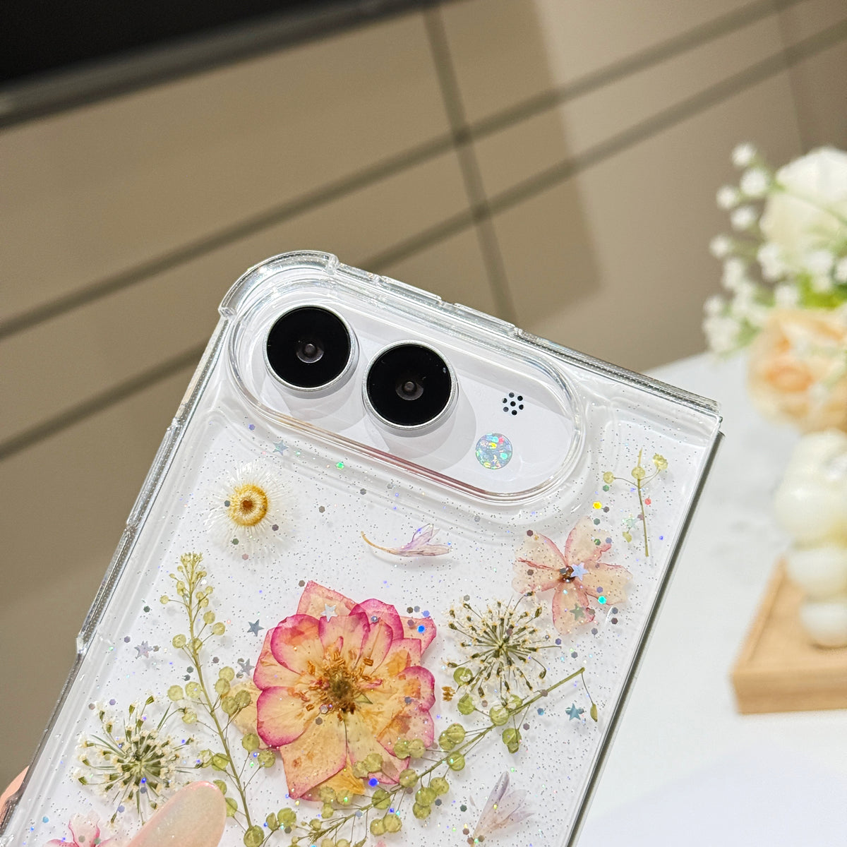 Close-up top-angle view of the clear iPhone DUO case embedded with real pressed dry flowers, fine botanical stems, and holographic star glitter surrounding the dual camera lens guard.