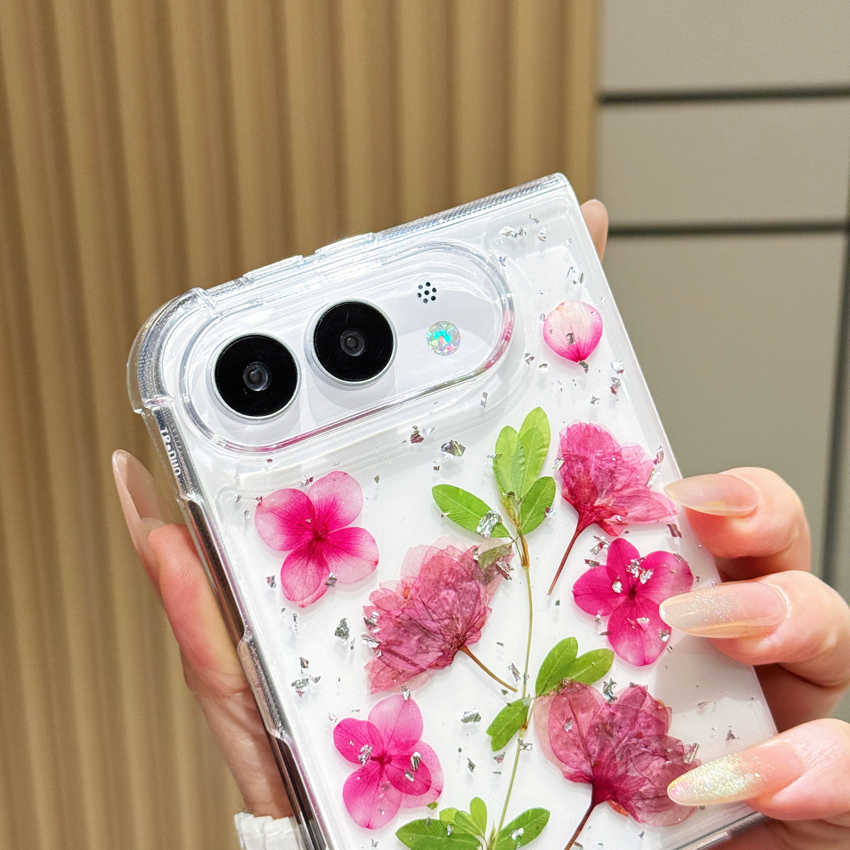 Close-up top-angle view of the clear iPhone DUO case embedded with real pressed pink hydrangeas, green leaves, and silver foil flakes surrounding the dual camera lens guard.