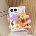 Close-up top-angle view of the clear iPhone DUO case embedded with real pressed pink tulips, purple wildflowers, green fern fronds, and gold foil flakes surrounding the dual camera lens guard.