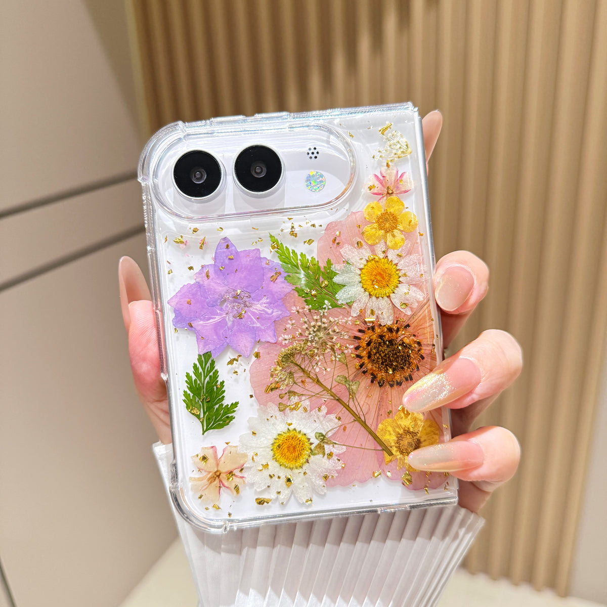 Close-up top-angle view of the clear iPhone DUO case embedded with real pressed pink tulips, purple wildflowers, green fern fronds, and gold foil flakes surrounding the dual camera lens guard.