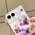 Close-up top-angle view of the clear iPhone DUO case embedded with real pressed purple hydrangeas, lilac petals, and shimmer glitter surrounding the dual camera cutout.