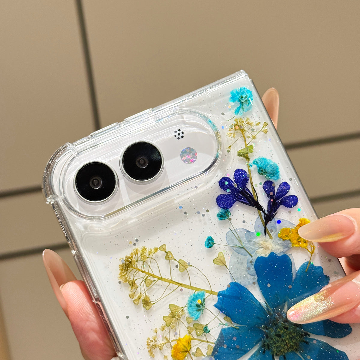 Close-up top-angle view of the clear iPhone DUO case showcasing the raised dual lens ring guard, tactile volume buttons, and embedded royal blue pressed flowers.