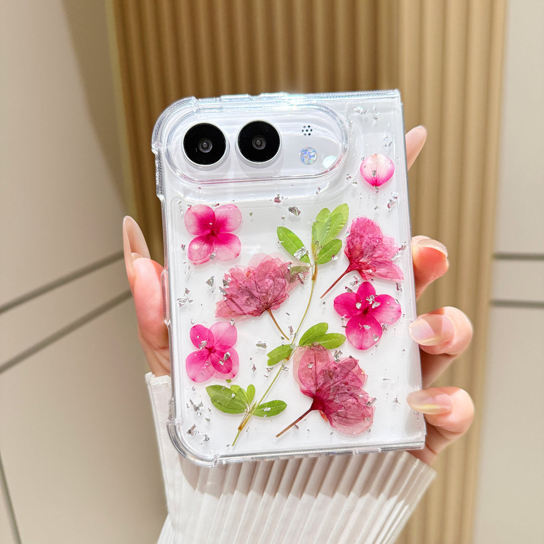 Close-up vertical perspective of a hand holding the clear iPhone DUO real pressed pink flower case, highlighting silver foil flecks and detailed leaf stems.