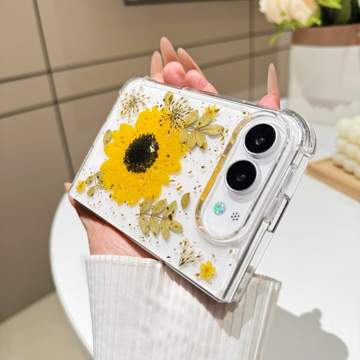 Close-up vertical perspective of a hand holding the clear iPhone DUO real pressed sunflower case, highlighting gold foil flecks and detailed botanical foliage layouts.