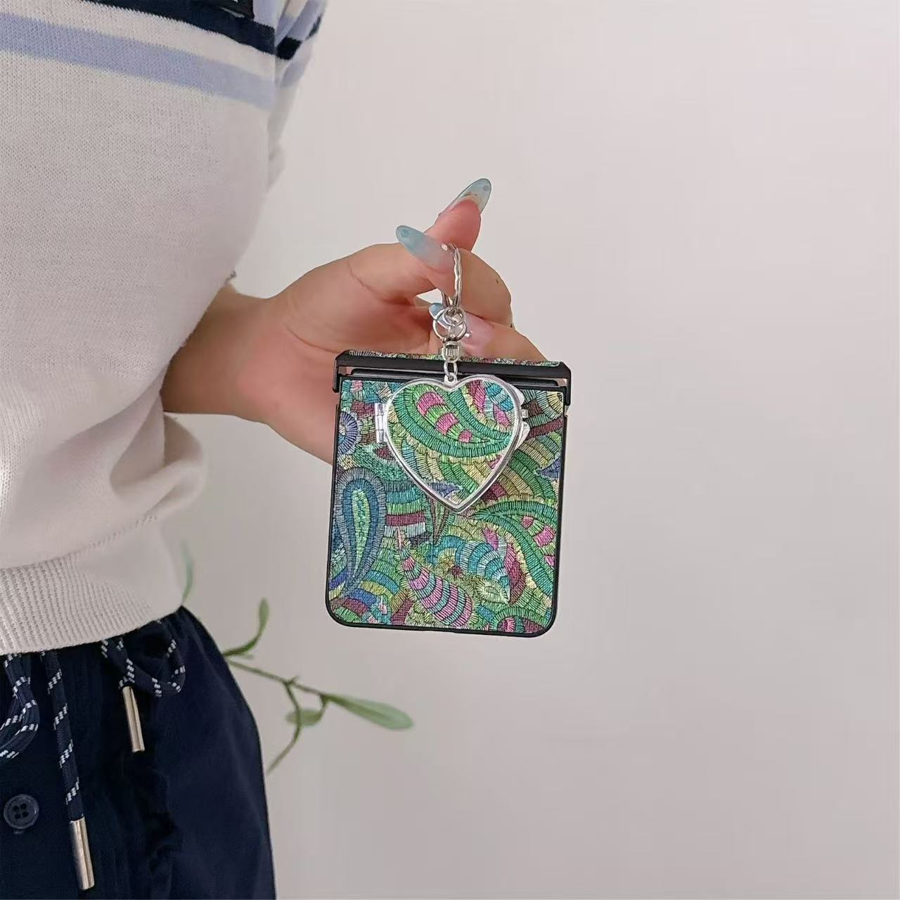 Close-up view of the green paisley fabric pattern phone case with heart compact mirror charm attached to top ring.