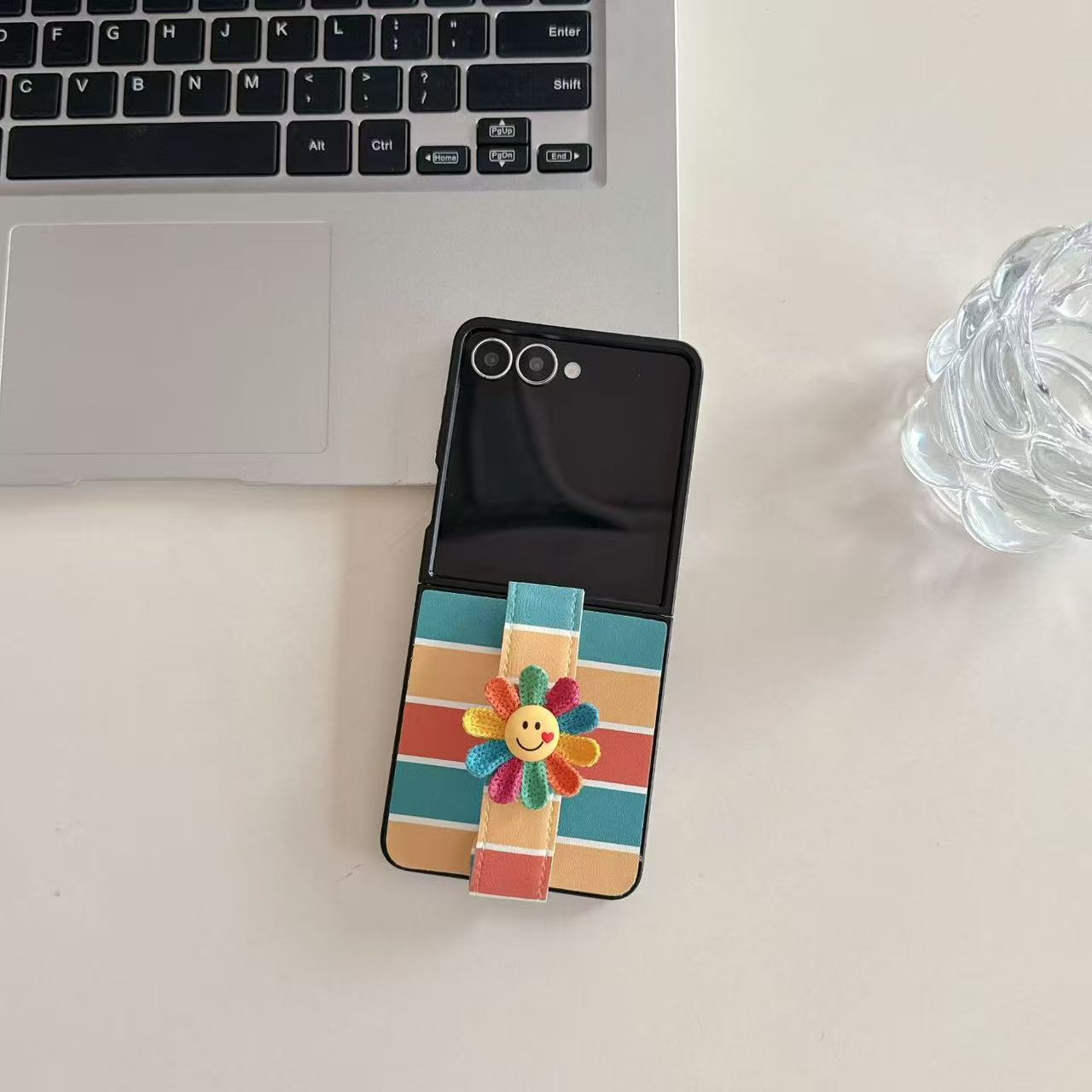 ⁠Colorful rainbow striped Samsung Galaxy Z Flip case lying open next to a laptop with a smiling flower hand strap.⁠