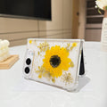 Desk display shot of the iPhone DUO clear case with a real pressed sunflower and gold flakes standing open on a white tabletop next to a white rose floral arrangement.