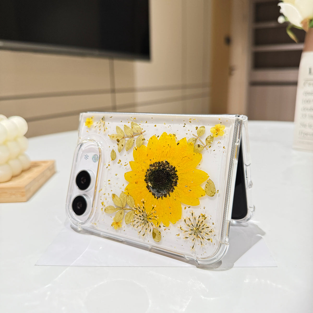 Desk display shot of the iPhone DUO clear case with a real pressed sunflower and gold flakes standing open on a white tabletop next to a white rose floral arrangement.