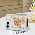 Desk display shot of the iPhone DUO clear case with real pressed dry flowers and star glitter standing open on a white tabletop next to a white rose floral arrangement.