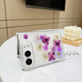Desk display shot of the iPhone DUO clear case with real pressed purple flowers and shimmer glitter standing open on a white tabletop next to a white candle setting.