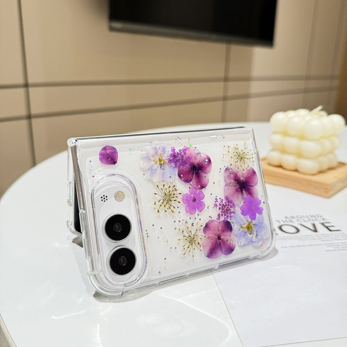 Desk display shot of the iPhone DUO clear case with real pressed purple flowers and shimmer glitter standing open on a white tabletop next to a white candle setting.