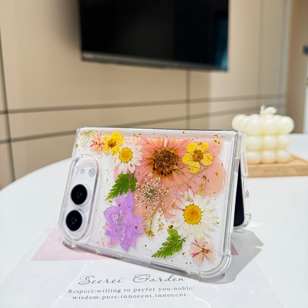 Desk display shot of the iPhone DUO clear case with real pressed tulip petals, wildflowers, and gold foil flakes standing open on a card next to a candle setting.