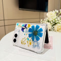 Elegant desk display of the open iPhone DUO clear case with real pressed blue and yellow garden flowers resting on a card next to a white rose floral arrangement.