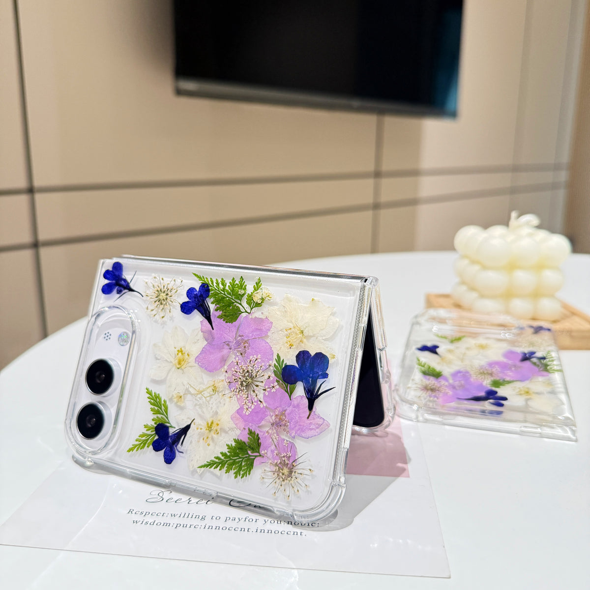 Elegant desk display of the open iPhone DUO clear case with real pressed garden flowers resting on a card next to a candle setting.