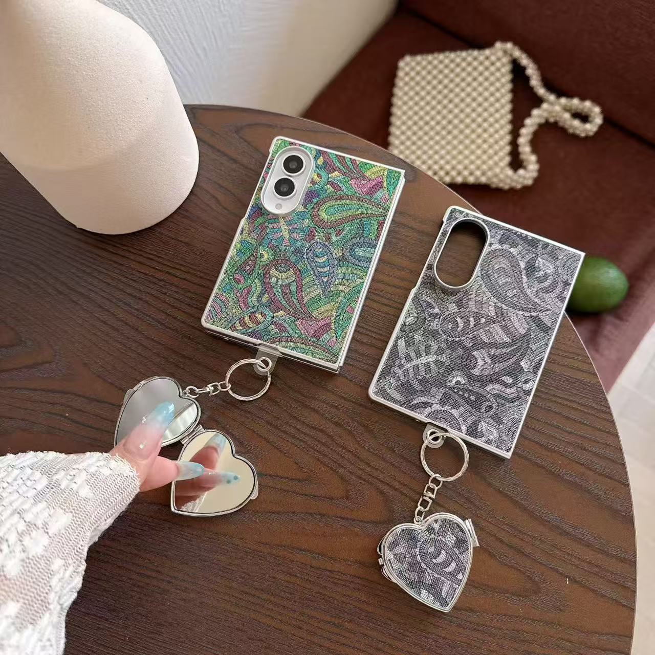 Green multi-color and black paisley tapestry Samsung Galaxy Z Fold 8 cases with open heart mirror charms on a dark wood table.