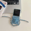 Flat lay photo of unfolded Samsung Z Flip denim floral case with wrist chain lying on a desk next to a laptop.
