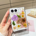 Flat lay product display of the clear iPhone DUO case featuring real pressed tulips, wildflowers, green leaves, and gold foil flecks on a white tabletop background.