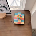 Folded retro rainbow stripe Galaxy Z Flip phone case displayed flat on a wooden desk with smiley flower embellishment.⁠