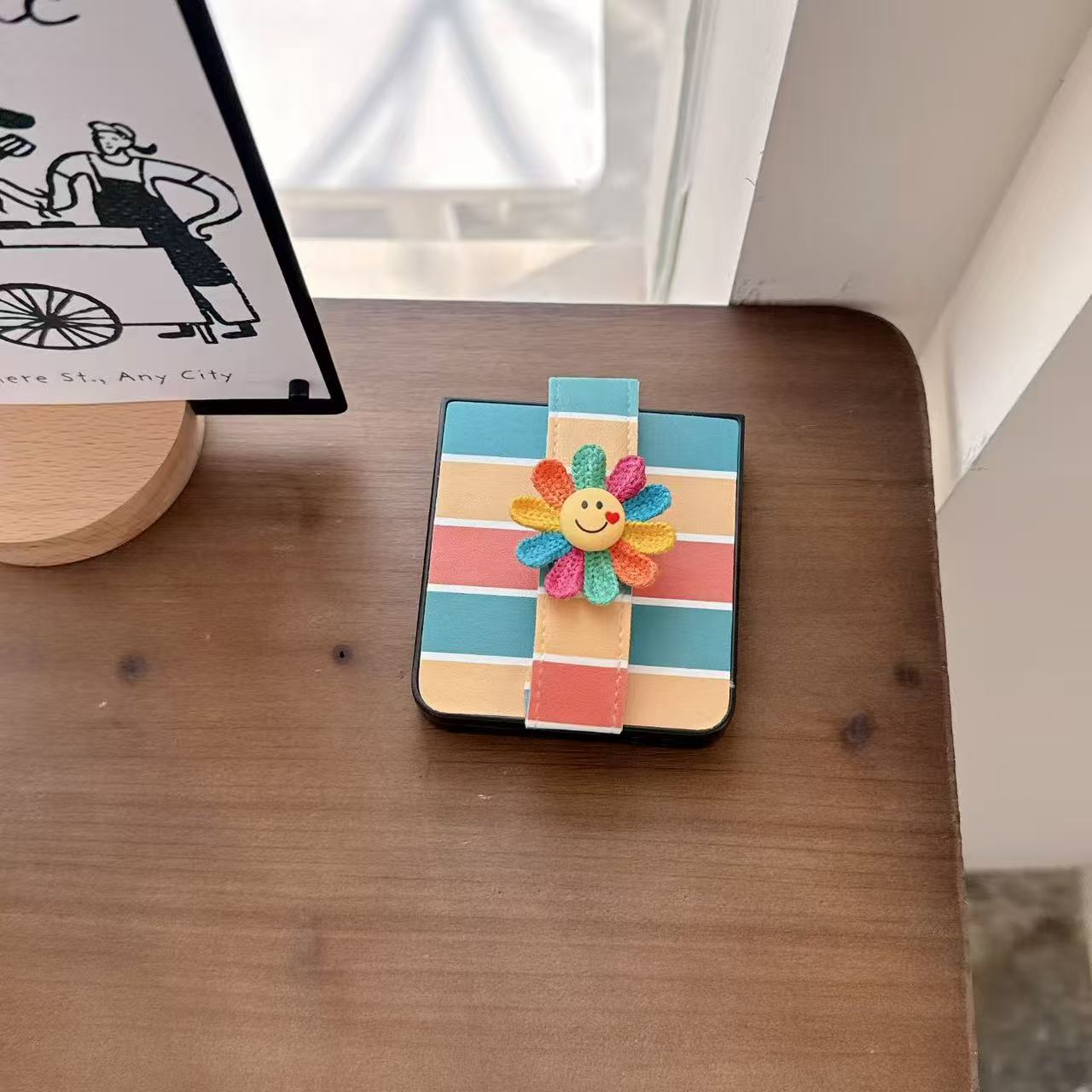 Folded retro rainbow stripe Galaxy Z Flip phone case displayed flat on a wooden desk with smiley flower embellishment.⁠