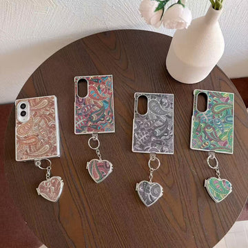 Green multi-color and black paisley tapestry Samsung Galaxy Z Fold 8 cases with open heart mirror charms on a dark wood table.