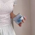Hand holding a folded Samsung Galaxy Z Flip case with a denim flower loop strap for secure grip.